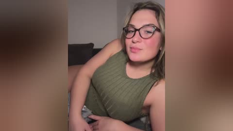 Video of a fair-skinned, plus-sized woman with blonde hair, wearing glasses, a green tank top, and a patterned skirt. She puckers her lips and looks directly at the camera, indoors with blurred background.
