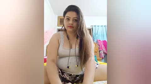 Video of a plus-sized, Latina woman with long brown hair, wearing a grey tank top and floral skirt, seated in a cluttered bedroom.