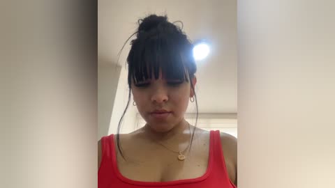 A young woman with medium skin tone and dark hair in a messy bun, wearing a red tank top, stands against a light-colored wall, slightly out of focus.