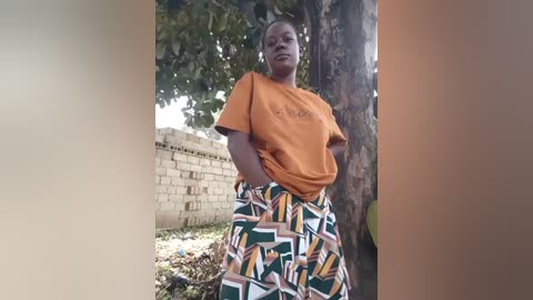 Video of a woman with medium brown skin, wearing an orange t-shirt and a patterned skirt, standing outdoors near a tree and a concrete wall, hands on hips, looking forward.