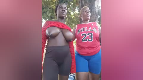 Video of two Black women, one topless with large breasts and the other in a red sports jersey, blue shorts, standing outdoors in a sunny, leafy background.