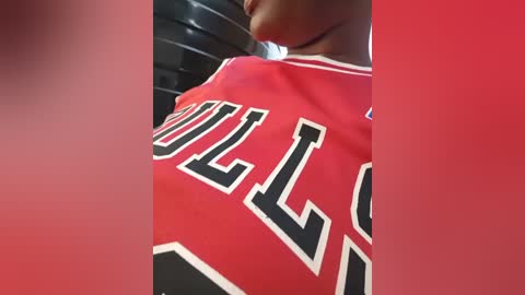 Video of a close-up of a person's chest in a red and black basketball jersey with the number \"1\" visible. The background features a blurred, dark-colored gym setting.