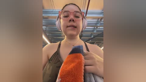 Video of a light-skinned woman with glasses, wearing a sleeveless olive-green top, holding an orange towel, in a modern, industrial setting with exposed beams and cables.