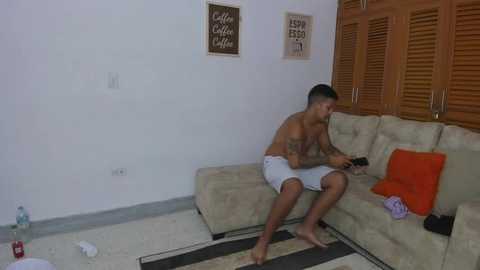 Video of a shirtless, muscular man in white shorts sitting on a beige couch, reading a book. Background features a white wall with wooden shutters and framed art.