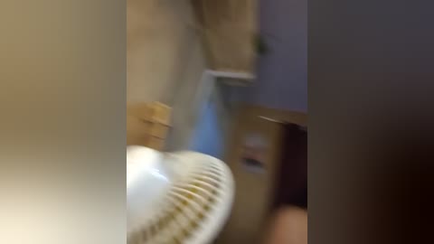 A blurry video of a beige wall, a wooden door, and a white oscillating fan in the foreground, with a person partially visible in the background.