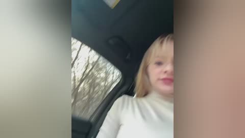 Video of a young woman with blonde hair and light skin, wearing a white top, seated in a car with a view of bare trees outside the window.