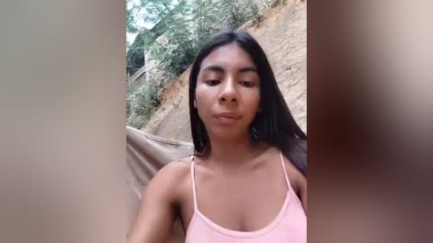 Video of a young Latina woman with medium brown skin and long black hair, wearing a pink spaghetti-strap top, standing outdoors against a cliffside background.
