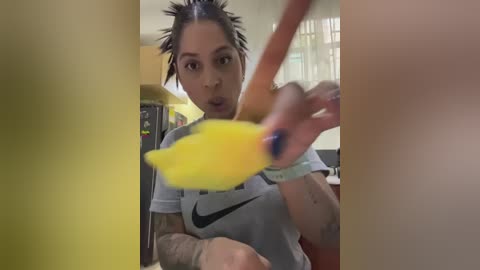 Video of a young woman with medium brown skin, black hair in braids, wearing a grey Nike T-shirt, holding a banana. Background includes a kitchen with cabinets and a yellow wall.