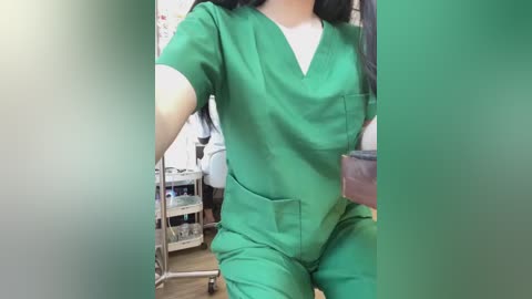 Media: Video of a person in green medical scrubs, wearing a white undershirt, holding a brown purse, standing in a clinical setting with medical equipment and white walls.