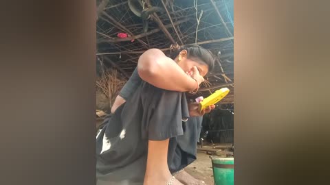 Video of a young Asian woman with medium build, wearing a black shirt and shorts, eating a yellow corn on cob in a rustic, open-air hut with bamboo walls.