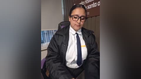 Video of a Latina woman with dark skin, wearing glasses, a black jacket, white shirt, and a navy tie, seated at a desk in an office with a blue curtain and a sign in Spanish.