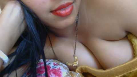 Video of a light-skinned woman with red lipstick, wearing a gold necklace, lying on a floral-patterned pillow, exposing ample cleavage, with long dark hair.