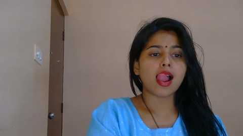 Video of a young South Asian woman with long black hair, fair skin, and a playful expression, sticking her tongue out. She wears a light blue t-shirt. Background shows a beige wall and a door.