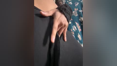 A video of a person's hand in motion, pulling black fabric. The background shows a teal floral-patterned shirt. The image is grainy and low-resolution, with a focus on the hand and fabric.