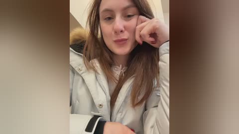 A video of a young woman with long brown hair, wearing a white coat, sitting indoors, looking pensive.