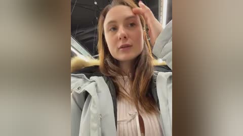 Video of a fair-skinned woman with long brown hair, wearing a light grey coat and pink sweater, adjusting her hair, inside a car with a black interior.