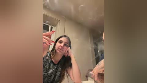 Video of a young woman with long dark hair, wearing a black and white patterned top, leaning on a bathroom counter, taking a selfie in a steamy bathroom.