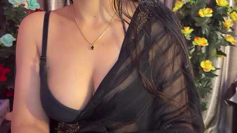 Video of a woman with fair skin, wearing a black sheer shawl over a black bra, showcasing medium-sized breasts. She has a gold necklace with a black pendant. Background features a floral arrangement with yellow and blue flowers.
