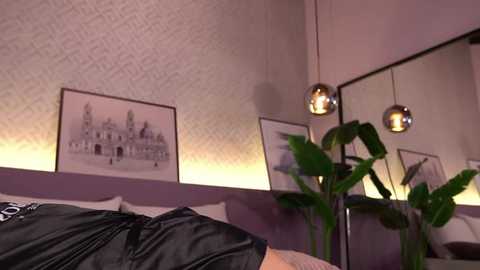 Video of a modern bedroom with a purple-patterned wallpaper, two pendant lights, a large potted plant, and a framed black-and-white architectural drawing. A person's arm and a black shirt are visible in the foreground.