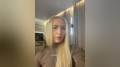 Video of a young, fair-skinned woman with long, straight blonde hair, wearing a brown turtleneck sweater, standing indoors with beige walls and modern decor.