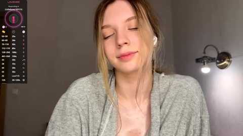 A video of a fair-skinned, blonde woman with closed eyes, wearing a gray cardigan, sitting indoors with a wall-mounted light fixture behind her.