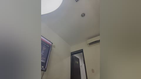 Video of a minimalist, modern room with off-white walls, a flat-screen TV mounted on the left, an air conditioning unit on the right, and a closed door.