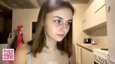 Video of a young woman with straight, shoulder-length brown hair, wearing a light blue tank top, standing in a kitchen. The setting includes white cabinets, a sink, and a hanging red coat.