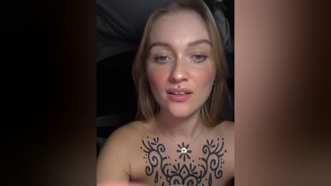 Video of a topless woman with light skin, blonde hair, and a floral tattoo on her chest, lying on a bed with dark sheets.