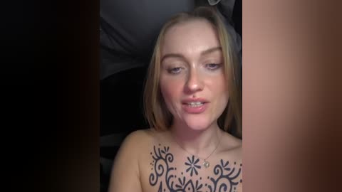 Video of a topless woman with light skin and blonde hair, wearing a necklace. She has intricate black tattoos covering her chest and shoulders. Background is blurred, focusing on her face.