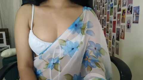 Media: Video of a woman in a white floral dress, showing off her cleavage, in a cluttered room with a collage of photos on the wall.