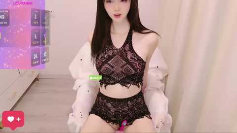 A video of a slender, fair-skinned woman with long, straight black hair, wearing a black lace lingerie set and white jacket, sitting on a bed in a light-colored room.