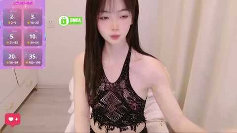 A video of an East Asian woman with long black hair, fair skin, and slender physique, wearing a black lace halter top, sitting on a bed in a minimalistic, beige room.