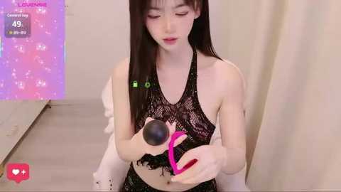 Video of a slender Asian woman with long black hair, wearing a black lace halter top and pink bra, using a pink vibrator. Background features a white curtain and a purple digital screen displaying her livestream.