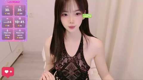 Video of a young East Asian woman with straight black hair, wearing a black lace halter top, in a minimalist room with white walls.