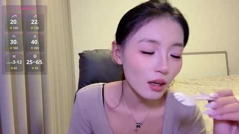 Video of an Asian woman with dark hair and light skin, wearing a beige top, eating with a white spoon, in a bed with beige sheets and a headboard.