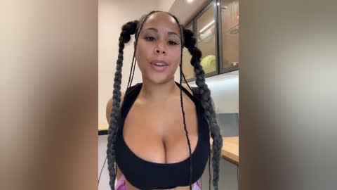 Video of a curvy Black woman with braided hair, wearing a black top that accentuates her large breasts, in a modern kitchen with stainless steel appliances.