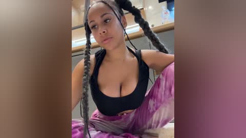 Video of a young Black woman with medium skin tone, large breasts, and long braided hair in a black sports bra and pink tie-dye pants. She sits in a modern kitchen with wooden cabinets and a white countertop.
