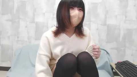 A video of an Asian woman with straight black hair and a white mask, wearing a beige sweater and black tights, sitting on a light blue chair against a white and gray patterned wall.