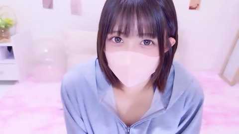 Video of a young Asian woman with straight black hair and light skin, wearing a light blue zip-up hoodie, sitting on a pink bed with a white headboard. She has a white face mask covering her nose and mouth.