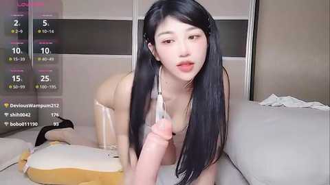 A video of an East Asian woman with long black hair, fair skin, and large breasts, wearing only a clear plastic dildo, holding it in front of her face, in a modern bedroom.