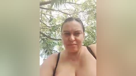 Video of a plus-sized, fair-skinned woman with auburn hair tied back, wearing a black bra, standing outdoors in a sunlit, leafy forest.