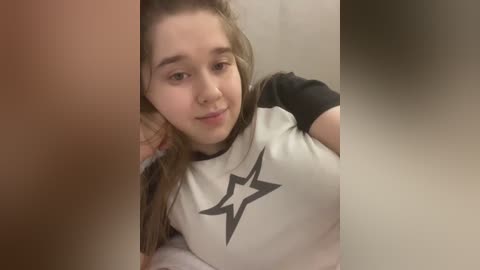 Video of a young woman with light skin, long brown hair, and a white T-shirt featuring a black star. She has a slight smile, and the background is blurred.
