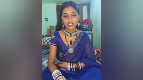 Video of a South Asian woman in a blue saree with intricate jewelry, sitting on a bed in a simple room with boxes and a wooden cabinet in the background.