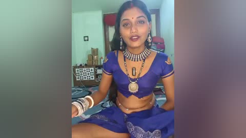 A video of a South Asian woman with long black hair, wearing traditional blue silk attire with intricate embroidery, multiple gold necklaces, and bangles. She sits in a modest, brightly lit room with patterned wall hangings and wooden furniture.