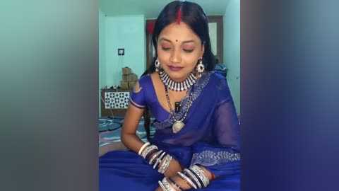 A video of a young Indian woman in a blue saree, adorned with traditional jewelry, including a red bindi, intricate earrings, and multiple bangles, seated in a simple room with white walls and a bed.