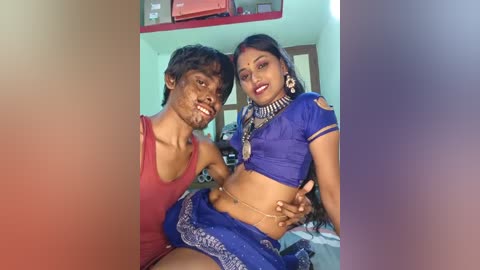 A video of a young South Asian couple in a casual indoor setting, the man wearing a red tank top, the woman in a blue sari.