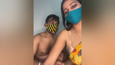 Video of a topless man with medium skin tone and a yellow and black striped mask, and a woman with a blue mask and yellow dress, in a dimly lit room.