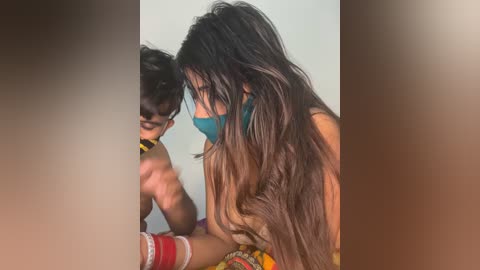 A video of a woman with long, wavy brown hair, wearing a teal face mask, applying makeup to a man with dark hair, both wearing colorful traditional attire, in a dimly lit room.