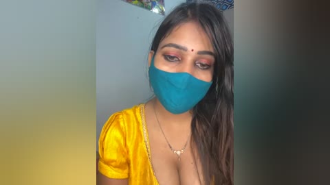 Video of a South Asian woman with long brown hair, wearing a yellow blouse, blue face mask, and red bindi, against a blurred, colorful background.