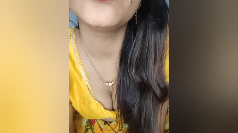 Video of a woman with long, dark hair, wearing a bright yellow kurta with floral patterns, and a gold necklace, partially visible cleavage.